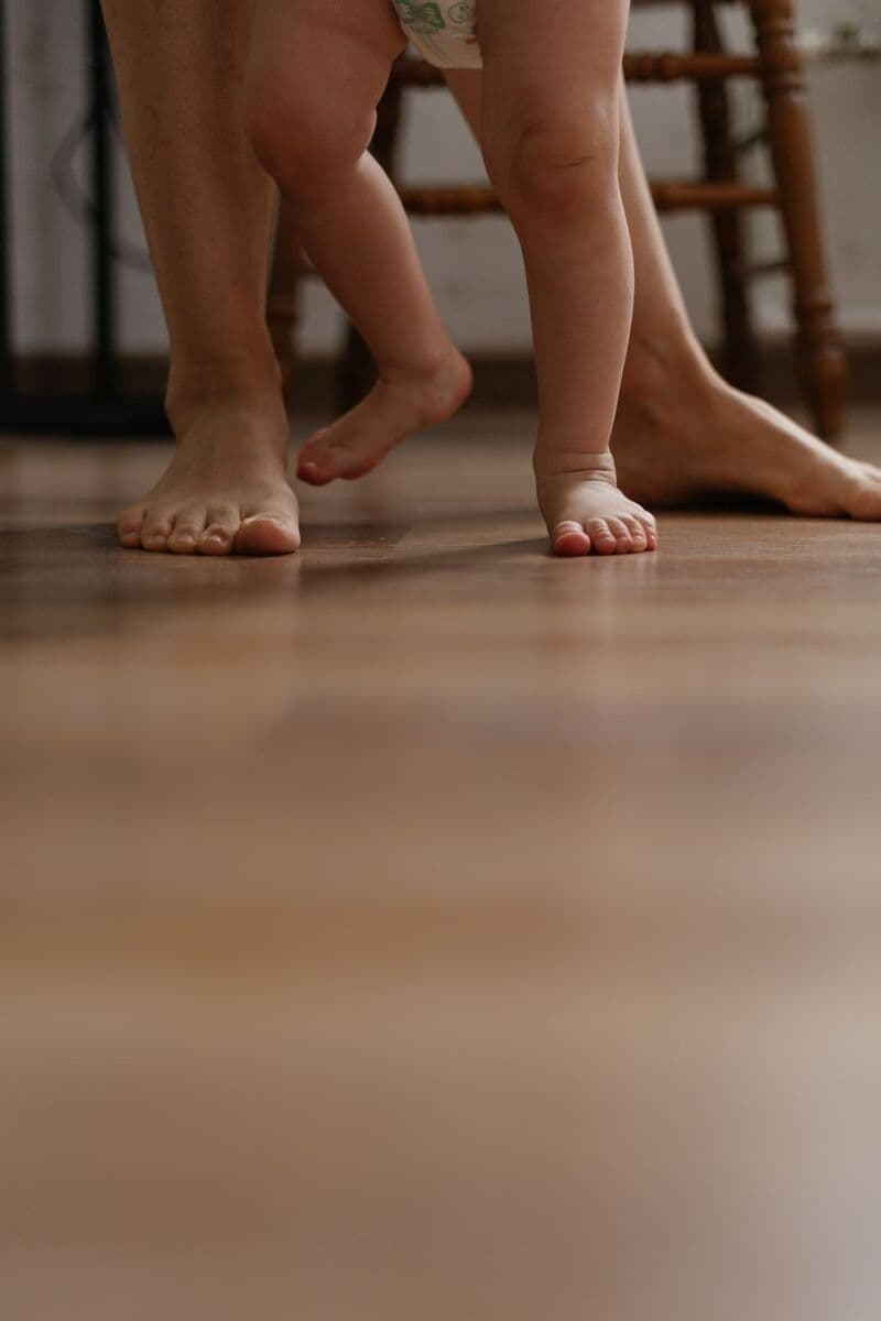 I piedi scalzi di un bambino che muove i primi passi su un pavimento di legno, accanto a quelli del genitore che lo sorregge.