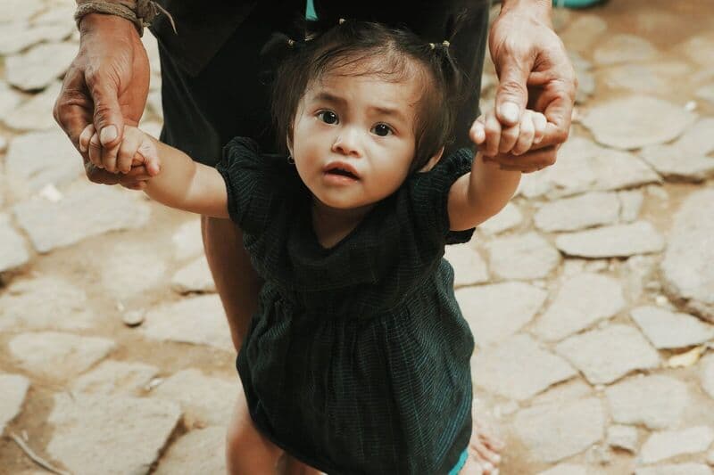 Una graziosa bambina muove i suoi primi passi all'aperto su un sentiero di pietra a Banten, in Indonesia, tenuta in braccio da un adulto.