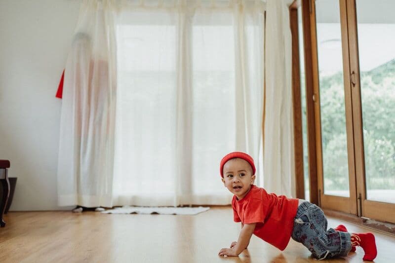 Un bambino carino vestito di rosso gattona su un pavimento in legno, giocando a nascondino in casa.
