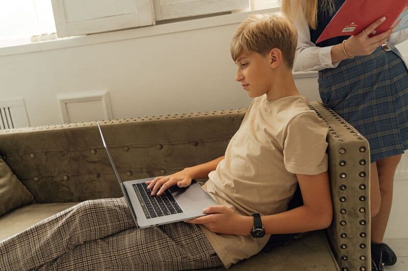 Un adolescente si rilassa su un divano con un laptop, impegnandosi nell'apprendimento online all'interno.