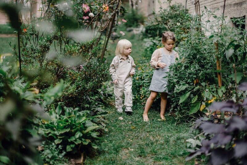 Due bambini esplorano un rigoglioso giardino domestico, circondati da una vegetazione vibrante e fiori.