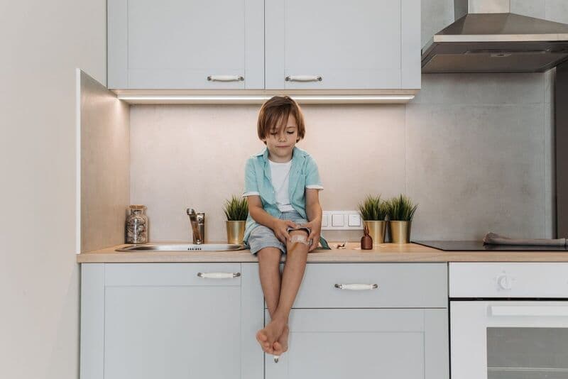 Bambino giovane seduto su un piano cucina moderno con il ginocchio fasciato, ambiente tranquillo.