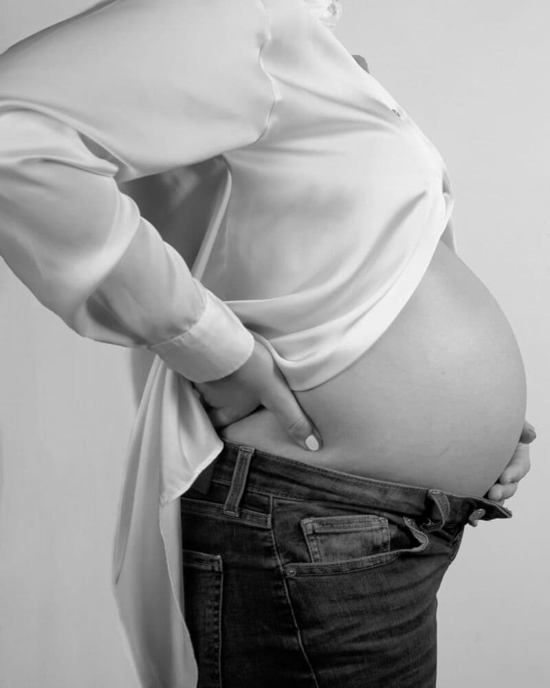Una donna incinta di profilo, in camicia bianca, con una mano appoggiata alla parte bassa della schiena.