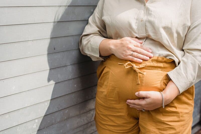 Primo piano di una donna incinta che accarezza il suo pancione contro un muro soleggiato.