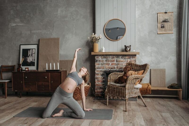 Donna incinta che pratica yoga vicino a un camino con un cane in una stanza accogliente.