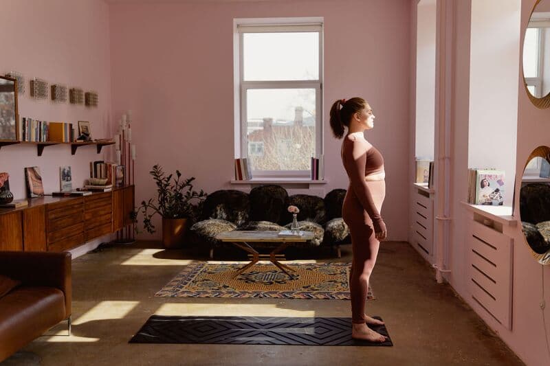 Una donna è in piedi su un tappetino da yoga all'interno, immersa nella luce naturale di una finestra, mentre pratica yoga.