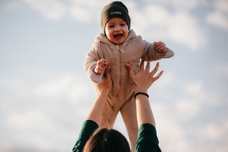 Un bambino sorridente con un berretto viene sollevato giocosamente verso il cielo in una giornata di sole.