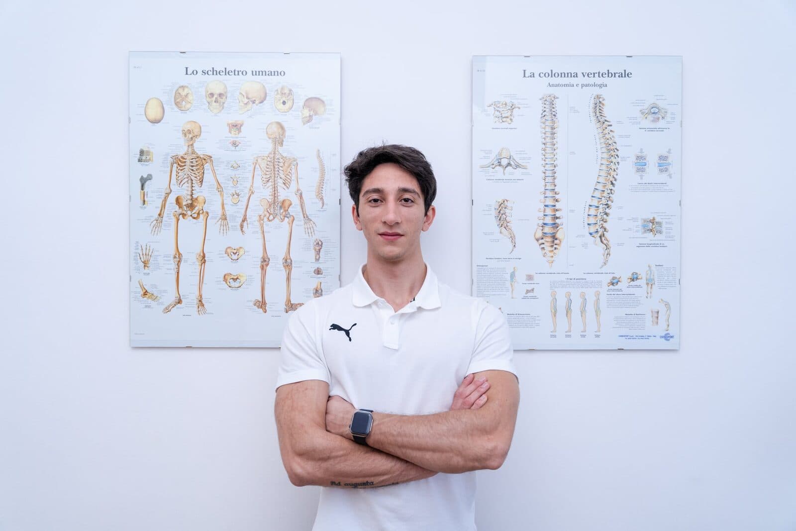 Fisioterapista in polo bianca con le braccia incrociate davanti a una parete bianca con due poster anatomici dello scheletro umano e della colonna ver