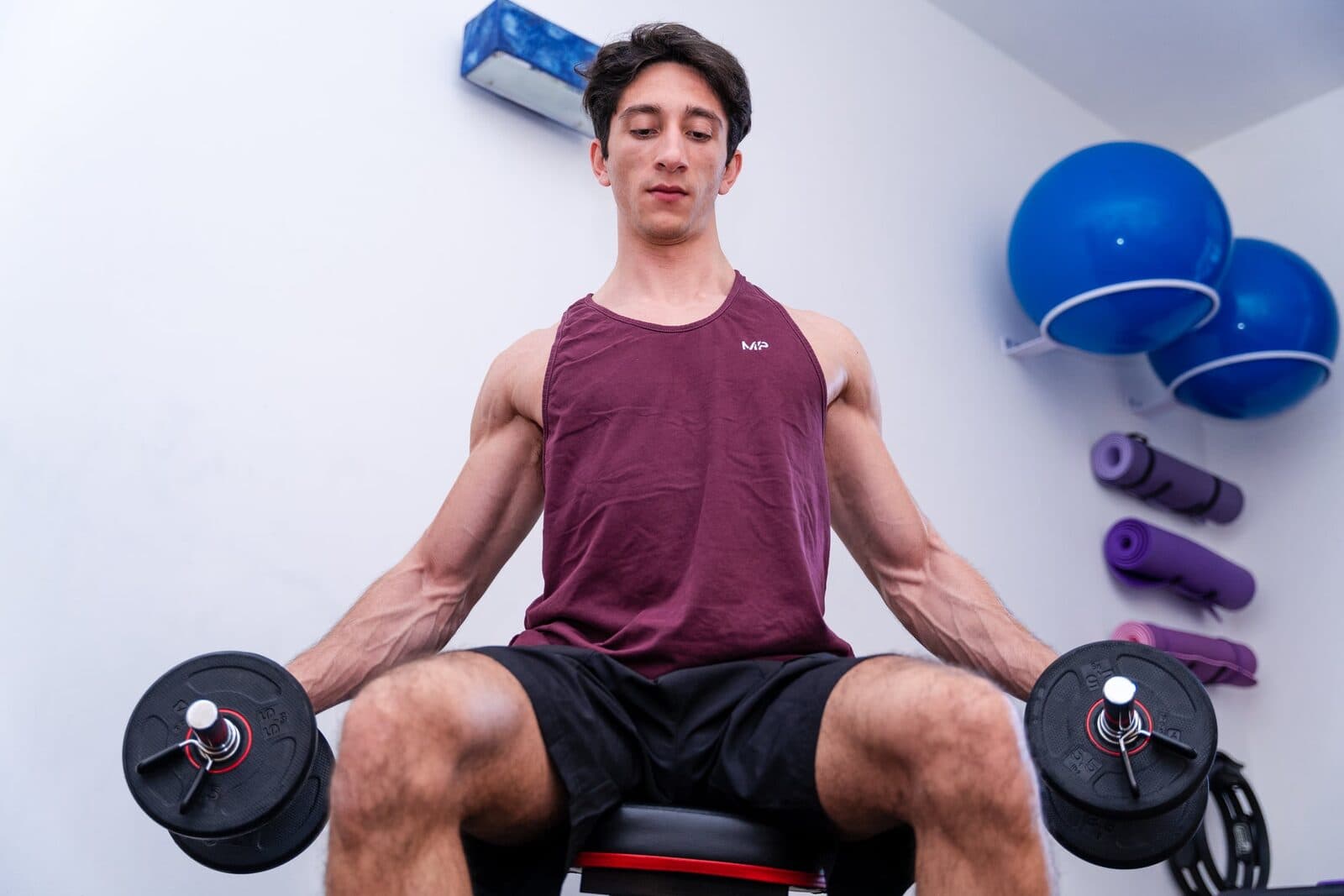 Uomo seduto con manubri lungo i fianchi, testa leggermente reclinata e occhi socchiusi nello sforzo, fitball blu e tappetini appesi dietro