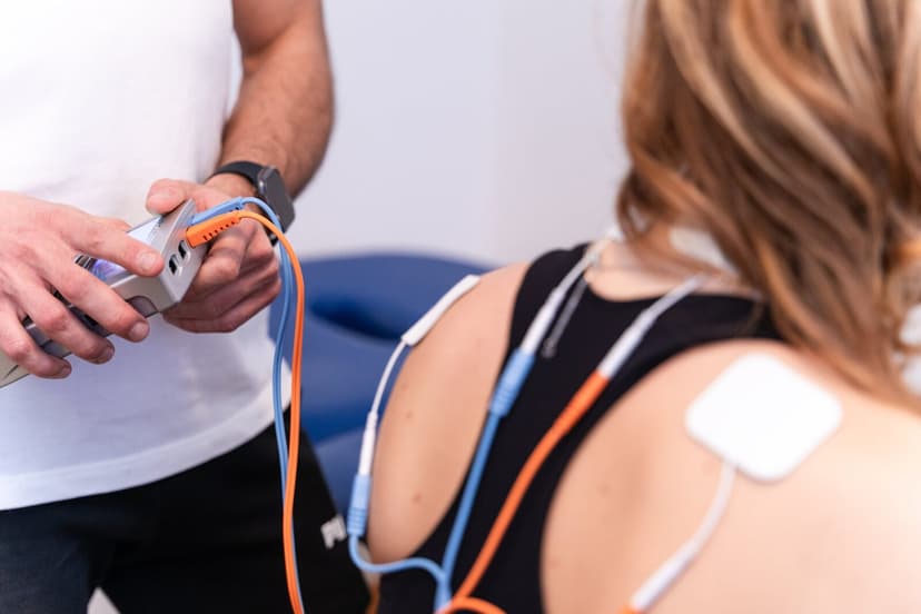 Le mani del fisioterapista regolano l'elettrostimolatore bianco tenendo il fascio di cavi arancioni; a destra, fuori fuoco, la schiena della paziente