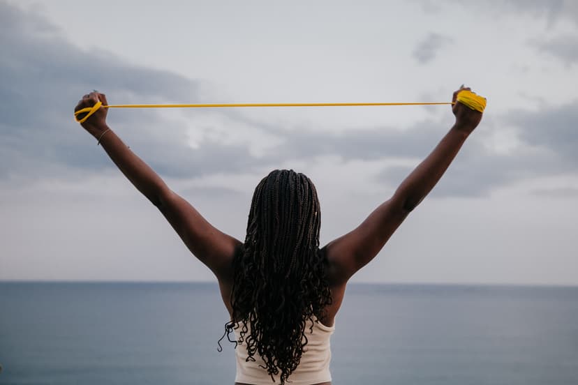 Una donna di spalle, davanti al mare, tende un elastico giallo sopra la testa tenendo le braccia aperte.