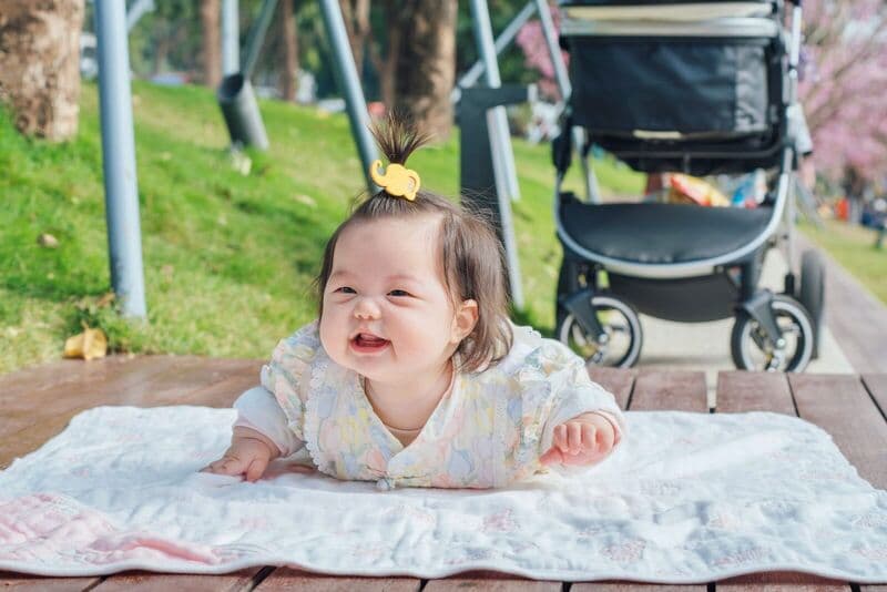 Un neonato asiatico sorridente è sdraiato su una coperta all'aperto, accanto a un passeggino, in una giornata di sole.