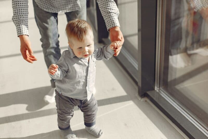 Un bambino che impara a camminare con il supporto di un genitore in uno spazio interno illuminato dal sole.