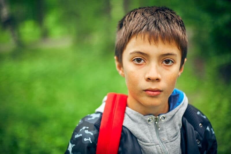 Un giovane ragazzo con uno zaino rosso si trova all'aperto circondato da vegetazione, catturato in un ritratto ravvicinato.