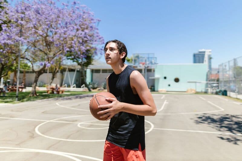 Adolescente in canotta nera con un pallone da basket su un campo all'aperto sotto un cielo azzurro e sereno.