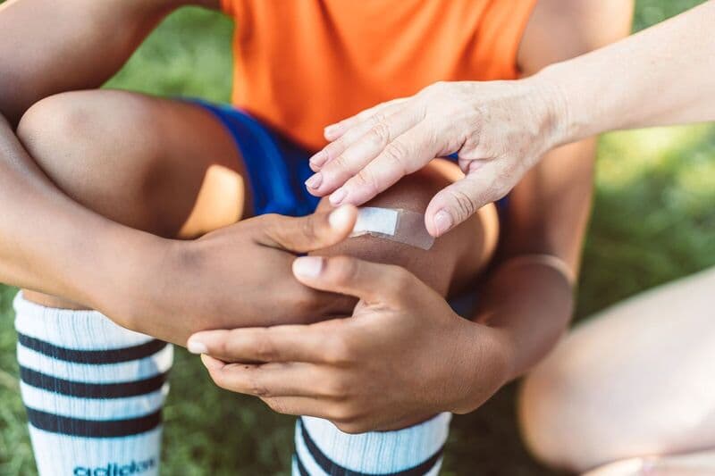Una vista ravvicinata di un adulto che applica un cerotto al ginocchio ferito di un bambino all'aperto durante il giorno.