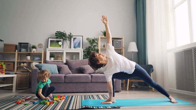 Una madre si allunga su un tappetino da yoga mentre il suo bambino gioca con dei blocchi vicino a loro nel soggiorno.