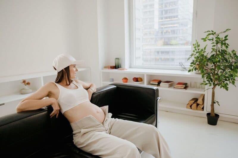 Donna incinta in abbigliamento casual si siede comodamente su un divano in una stanza moderna e ben illuminata.