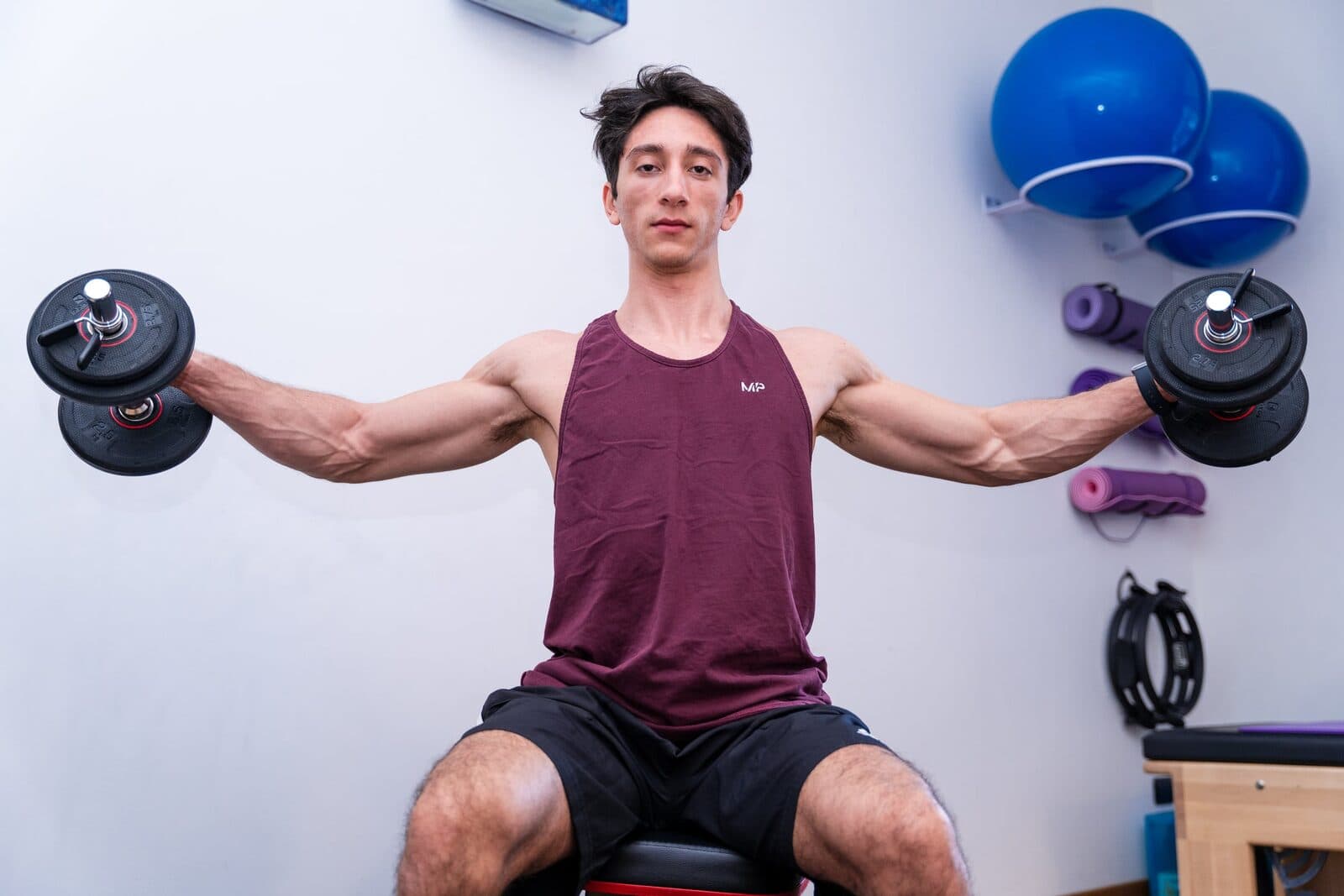 Alzate laterali con manubri: uomo in canotta bordeaux seduto su panca con le braccia distese all'orizzontale, sala attrezzi con fitball blu e tappetin