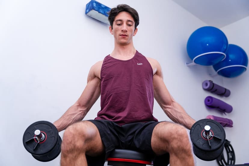 Uomo seduto con manubri lungo i fianchi, testa leggermente reclinata e occhi socchiusi nello sforzo, fitball blu e tappetini appesi dietro