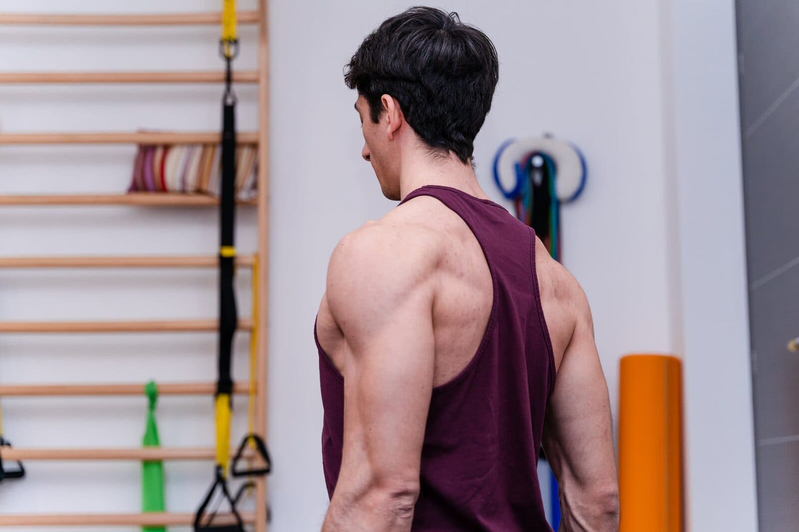 Uomo di spalle in canotta bordeaux, schiena e deltoidi in evidenza, davanti a una spalliera svedese con TRX ed elastici appesi