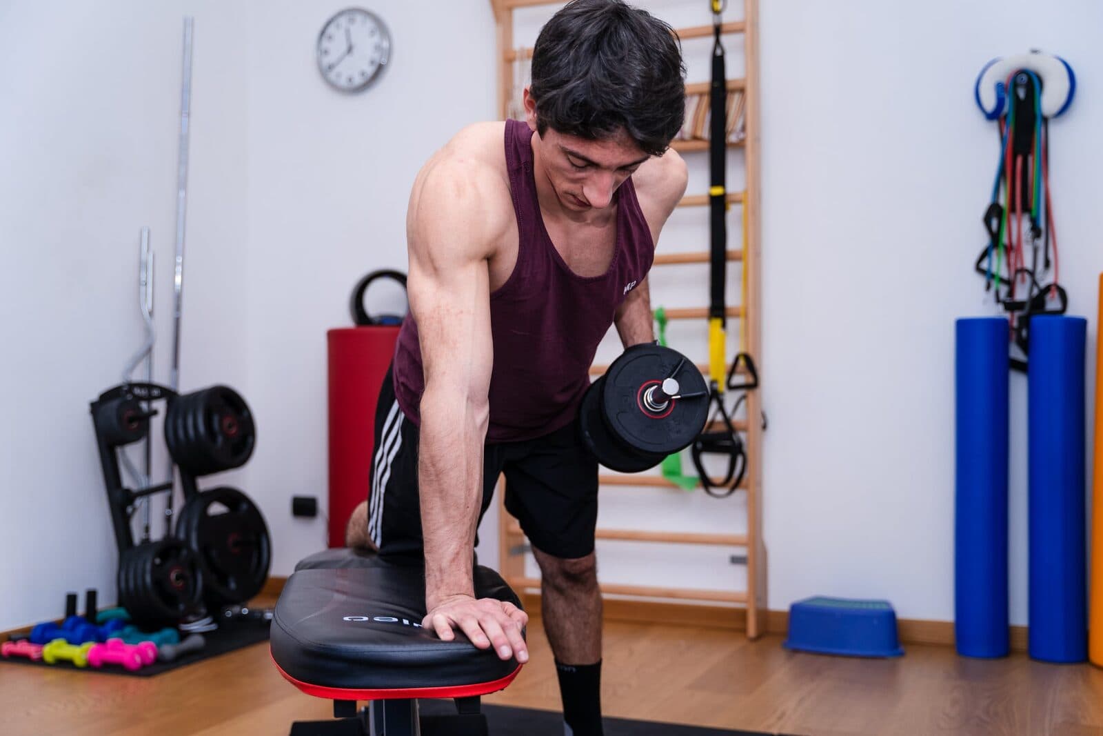 Uomo piegato in avanti sulla panca con il manubrio a metà tirata, testa leggermente ruotata, kettlebell e dischi sullo sfondo