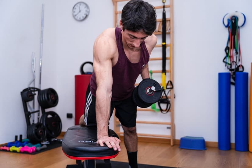 Uomo piegato in avanti sulla panca con il manubrio a metà tirata, testa leggermente ruotata, kettlebell e dischi sullo sfondo