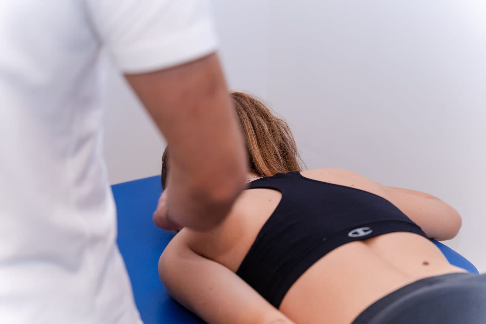 Donna prona sul lettino blu con top sportivo nero mentre le mani del fisioterapista in camice bianco lavorano sulla zona lombare