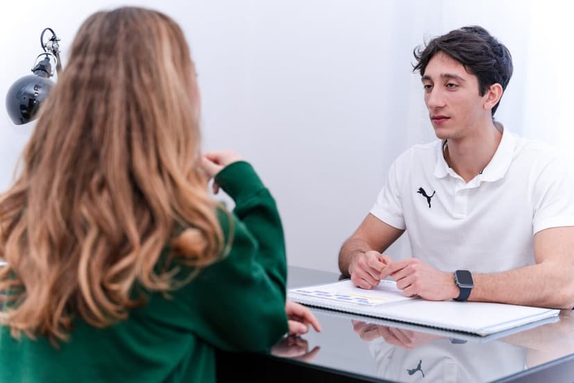 Inquadratura più larga del colloquio: la paziente di spalle in primo piano gesticola verso la propria spalla, Simone dall'altra parte del tavolo ascol