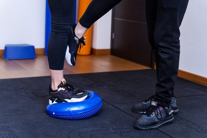 Dettaglio dei piedi: una scarpa in appoggio sulla cupola blu del bosu e l'altra gamba sollevata, accanto le scarpe del fisioterapista sul tappetino ne