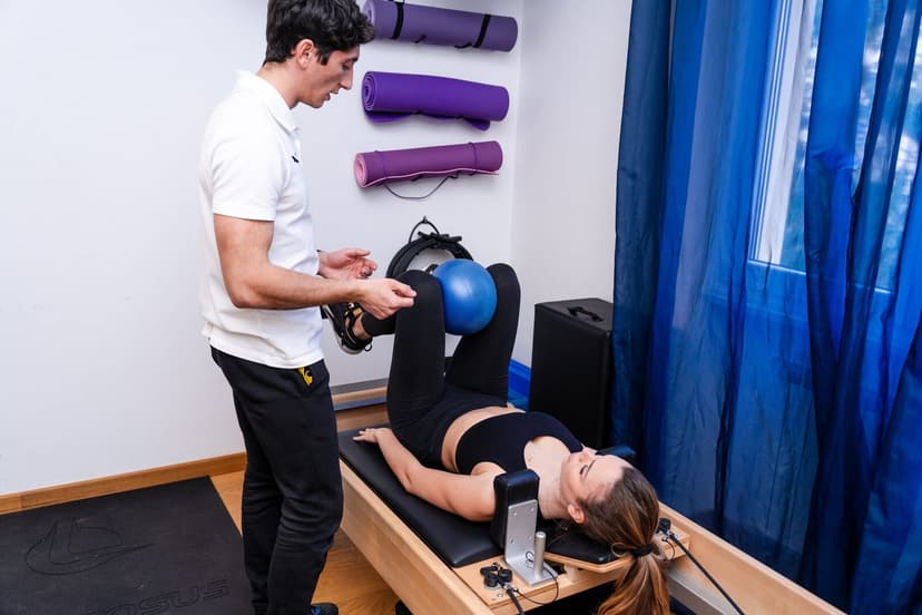 Campo largo dello studio: fisioterapista in piedi accanto al reformer che assiste la paziente distesa con palla blu tra le ginocchia, tappetini viola