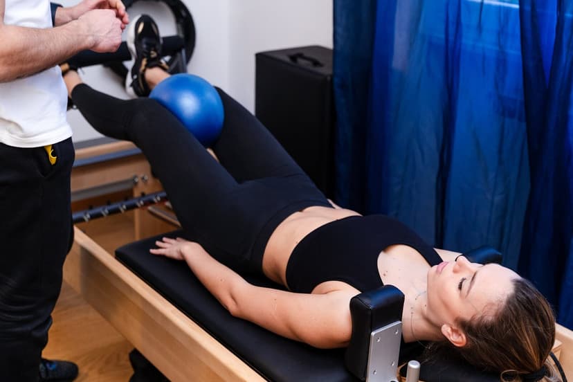 Vista laterale della paziente distesa sul reformer con gambe piegate e palla blu tra le ginocchia, braccia lungo il corpo; il fisioterapista compare s
