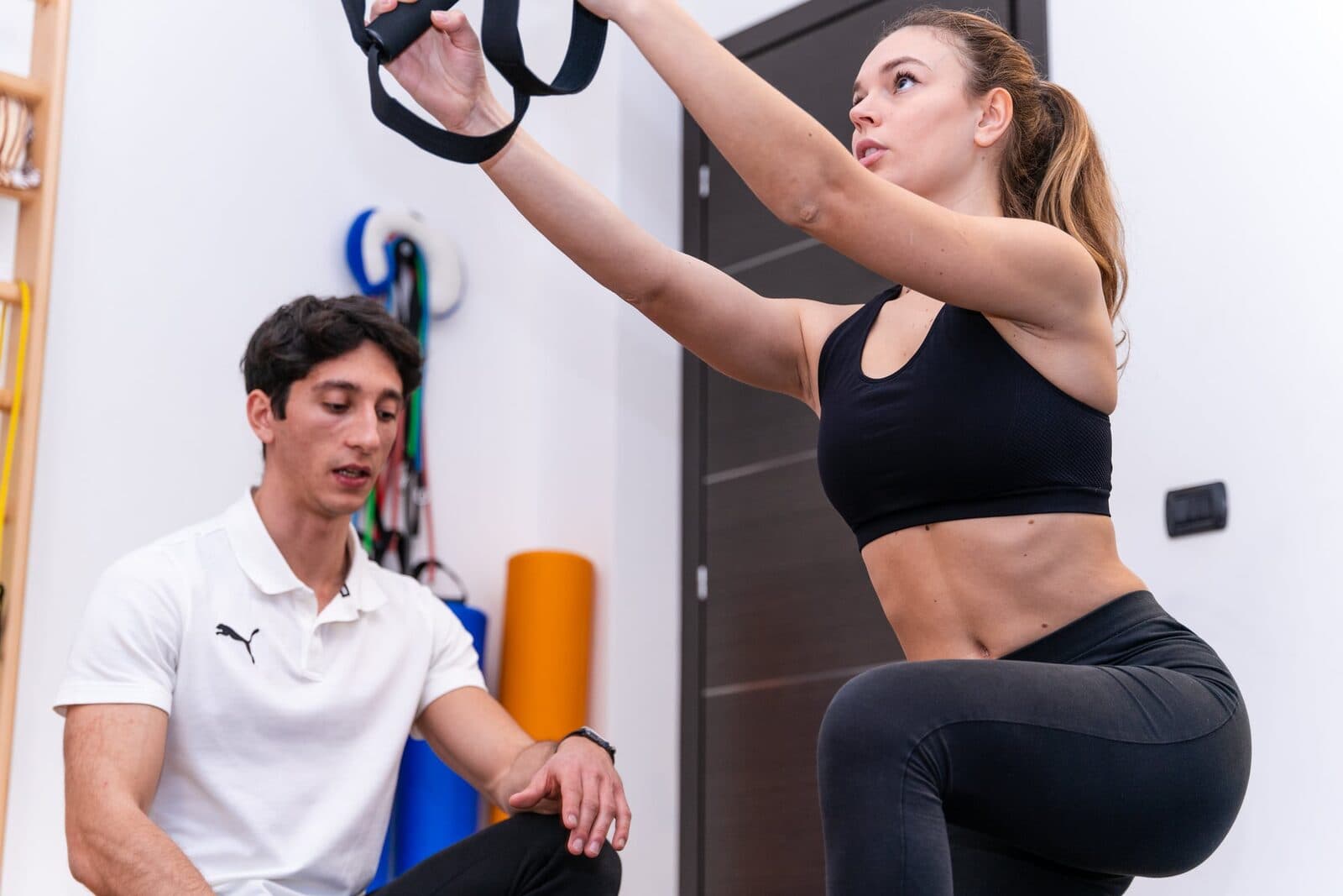 Ragazza con braccia distese in alto sul TRX vista di tre quarti, il fisioterapista seduto sui talloni la osserva da sotto; porta scura e spalliera sul