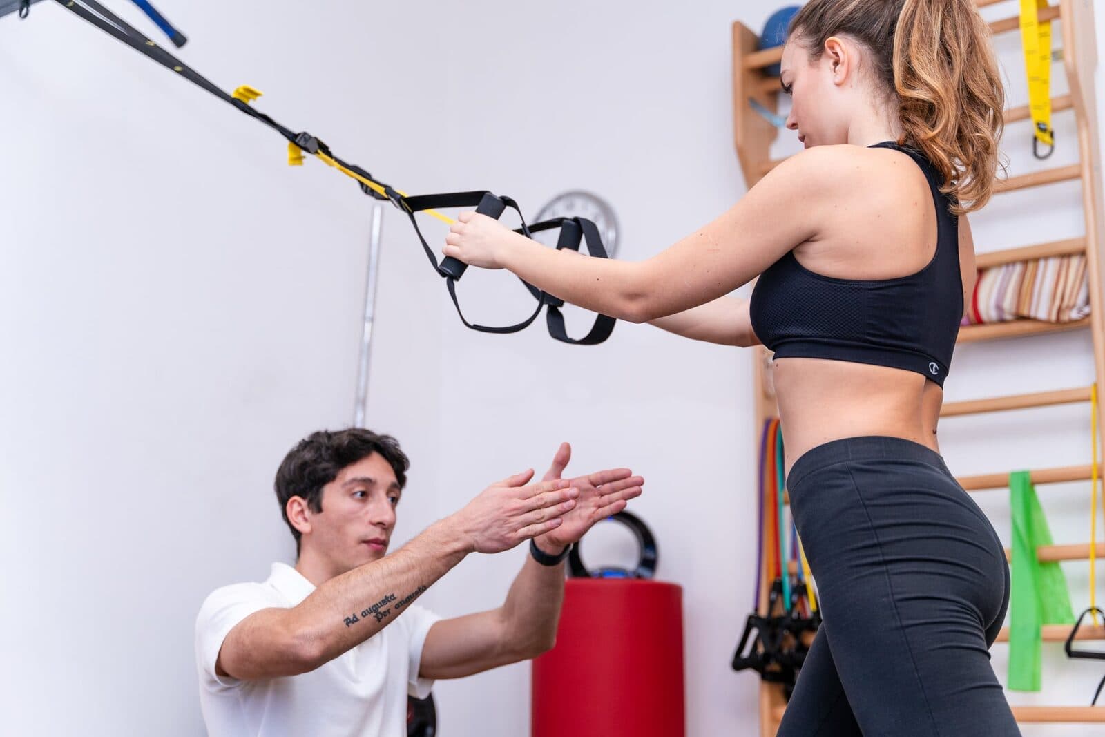 Fisioterapista accosciato con entrambe le mani protese che mima il movimento, mentre la ragazza in piedi tiene le braccia distese sul TRX giallo.