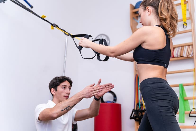 Fisioterapista accosciato con entrambe le mani protese che mima il movimento, mentre la ragazza in piedi tiene le braccia distese sul TRX giallo.