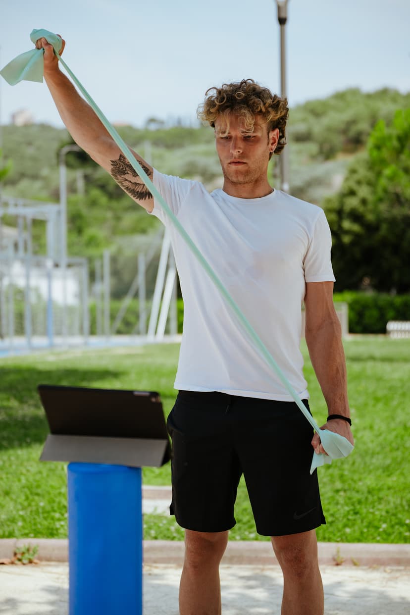Un ragazzo con la maglietta bianca tende un elastico verde sopra la testa, in un parco.