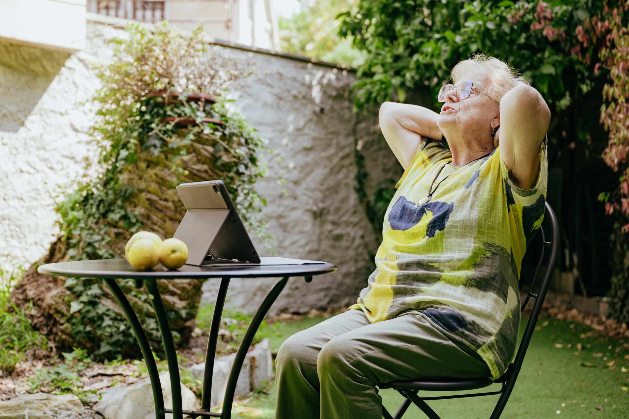 Una signora anziana seduta al tavolino del giardino si allunga con le mani dietro la testa, davanti al tablet appoggiato sul tavolo.