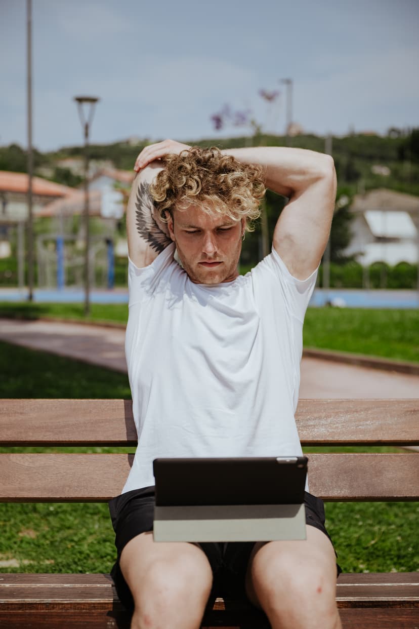 Ragazzo riccio seduto su una panchina di legno in un parco con un tablet appoggiato sulle gambe.