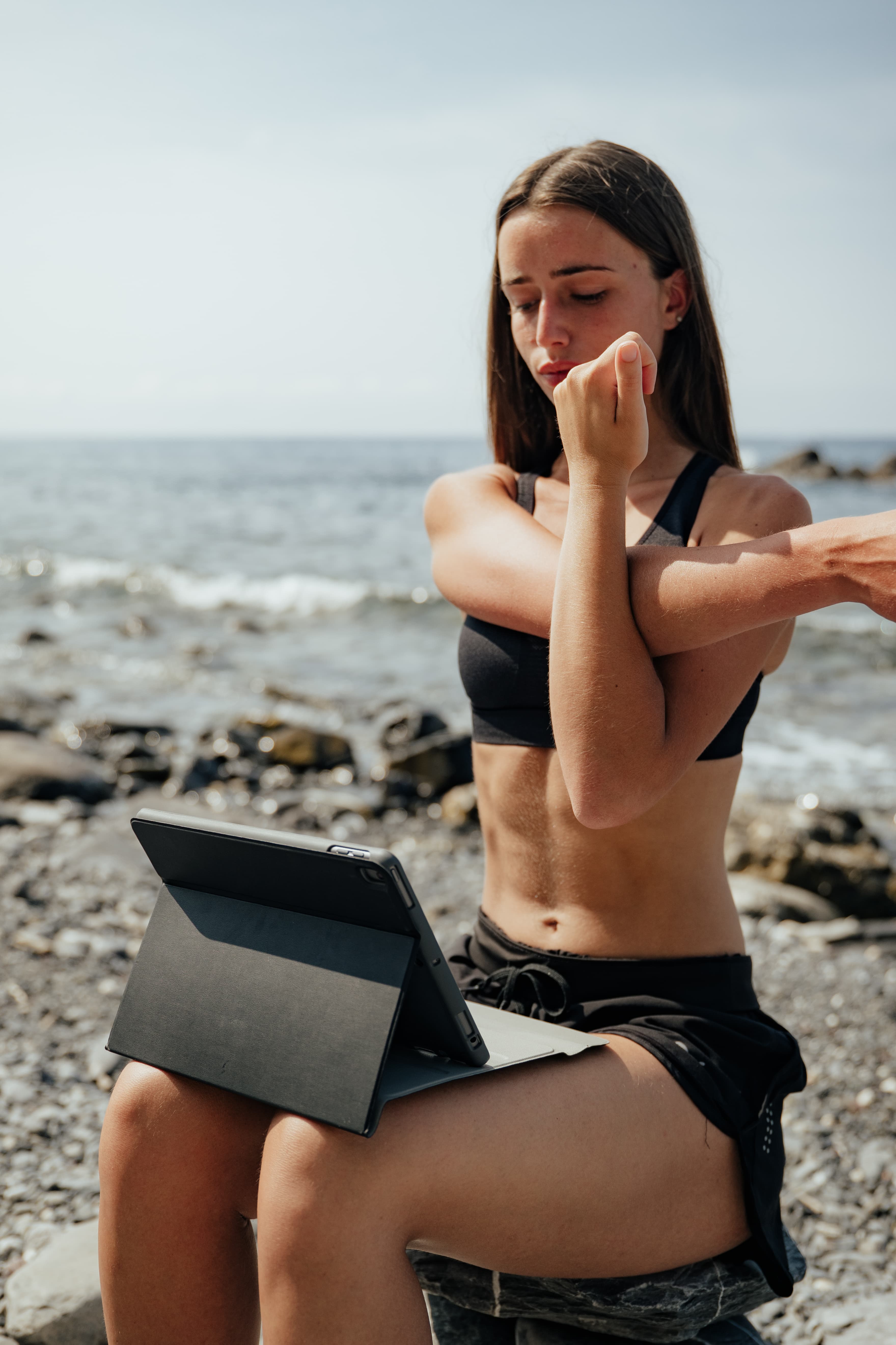 Una giovane donna seduta in riva al mare porta un braccio al petto per allungare la spalla, con il tablet appoggiato al ginocchio aperto sulla seduta.