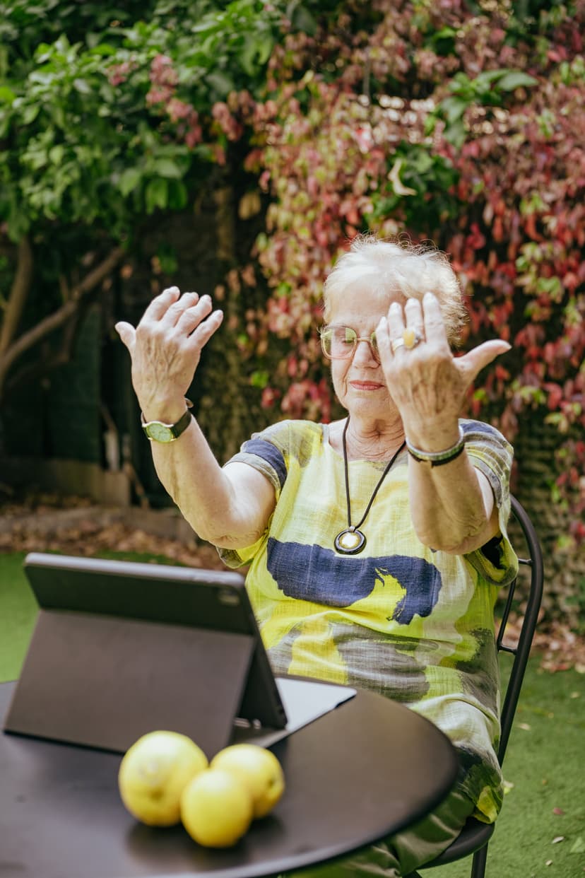 Signora anziana con capelli bianchi seduta a un tavolino da giardino che solleva le mani davanti a un tablet.